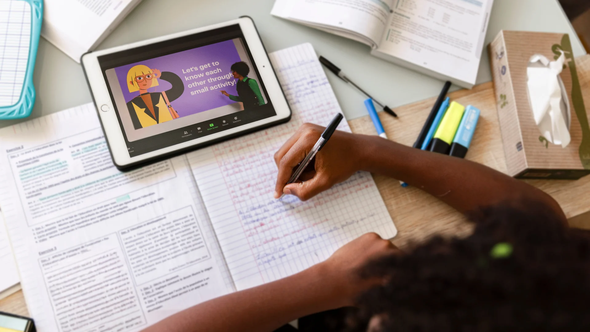 Child attending an online class on a tablet while writing notes in a notebook, surrounded by textbooks, pens, and highlighters on a study desk