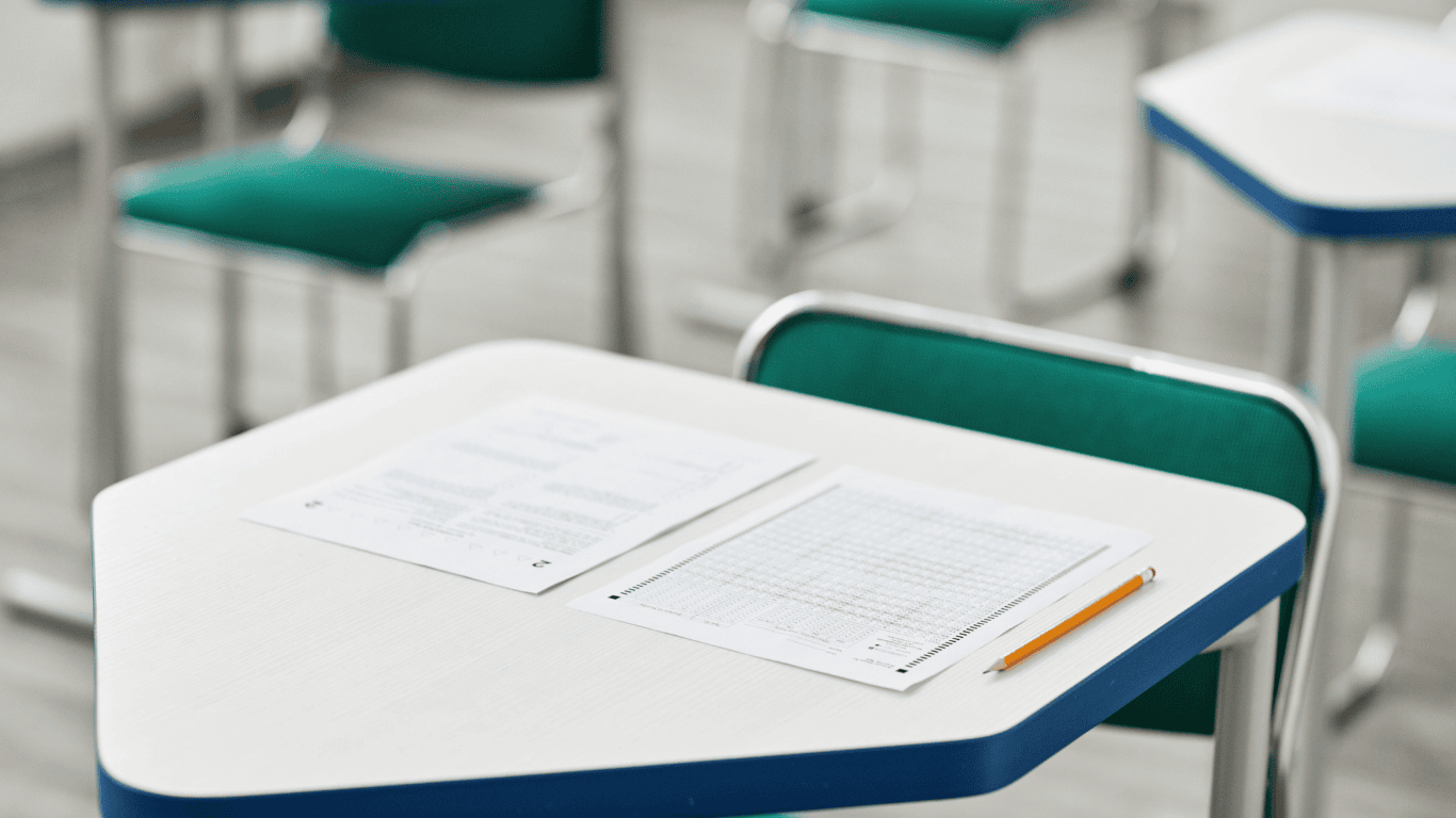 A exam hall with multiple desks, one of which has an exam sheet, an answer sheet, and a sharpened yellow pencil placed on it. The desks have green chairs, and the setting appears to be prepared for a test.