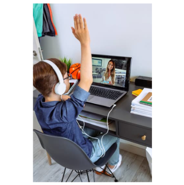 A young boy wearing headphones raises his hand while attending an online class on his laptop. He sits at a study desk with books, a basketball, and school supplies in the background.