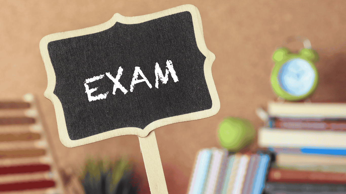A small chalkboard sign with the word 'EXAM' written on it, placed on a study desk with blurred books, a clock, and other study materials in the background.