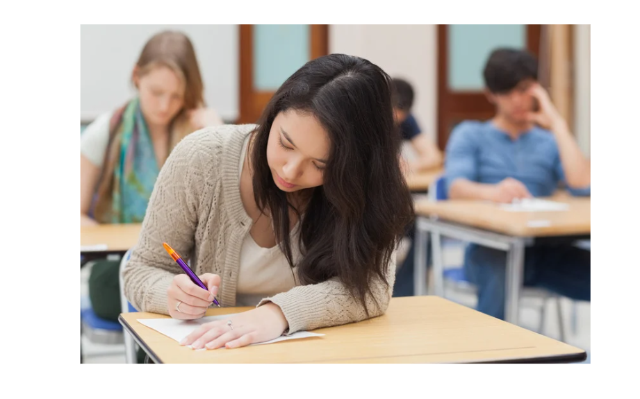 A young woman in a beige sweater writes attentively in a classroom, with other students in the background