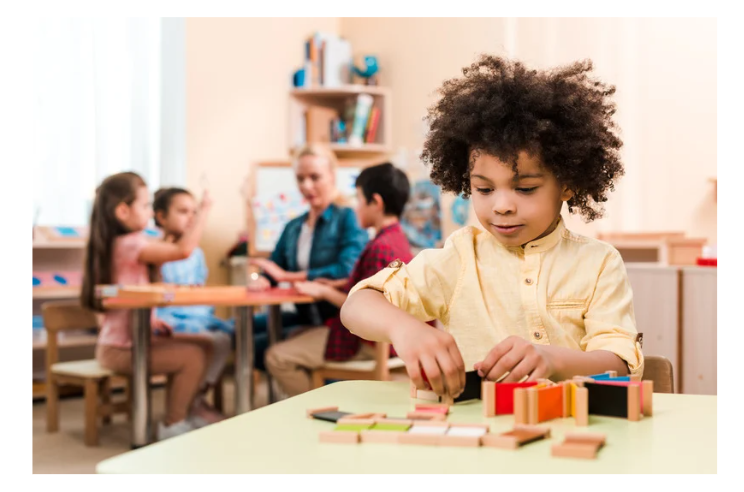 A young child engages in a hands-on activity with colorful wooden blocks at a Montessori classroom table, while other children and a teacher interact in the background.