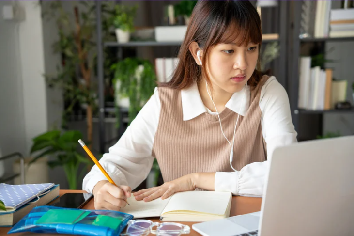 Young woman studying at a desk with a laptop, taking notes in a notebook while wearing earphones, in a well-lit and green indoor setting
